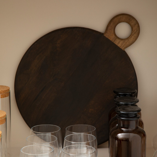 A dark wooden chopping board with loop handle on a shelf with some glasses in front of it