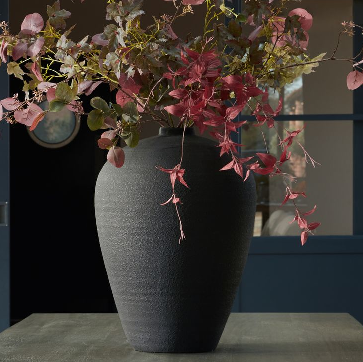 A large matt black vase with autumnal flowers in it, on a table