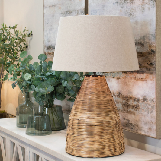 A table lamp with a woven wicker base and pale linen shade, on a side table with some vases